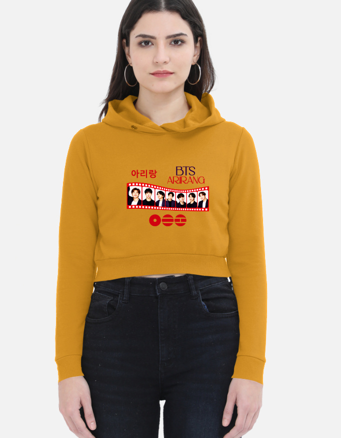Woman wearing a mustard yellow cropped hoodie featuring a BTS "Arirang" film strip graphic print on the front with member portraits and Korean Hangul text.